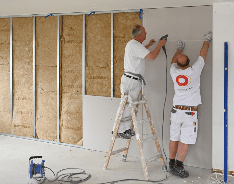 Zwei Handwerker: Einer steht auf einer Leiter und hält eine Bohrmaschine. Der zweite nebendran assistiert ihm und hält ein Massband an der Wand.
